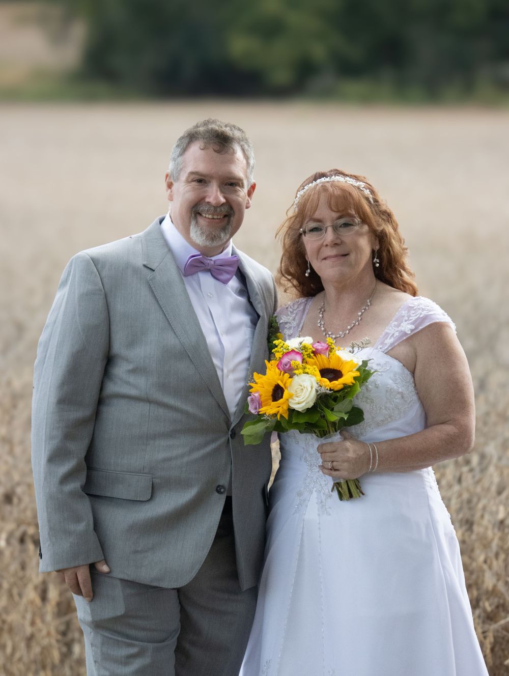 A couple dressed in wedding attire poses in a wheat field with sunflower bouquet.