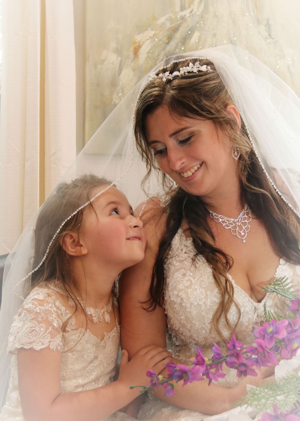 A bride in a white lace gown and veil shares a tender moment with a young attendant in matching lace.