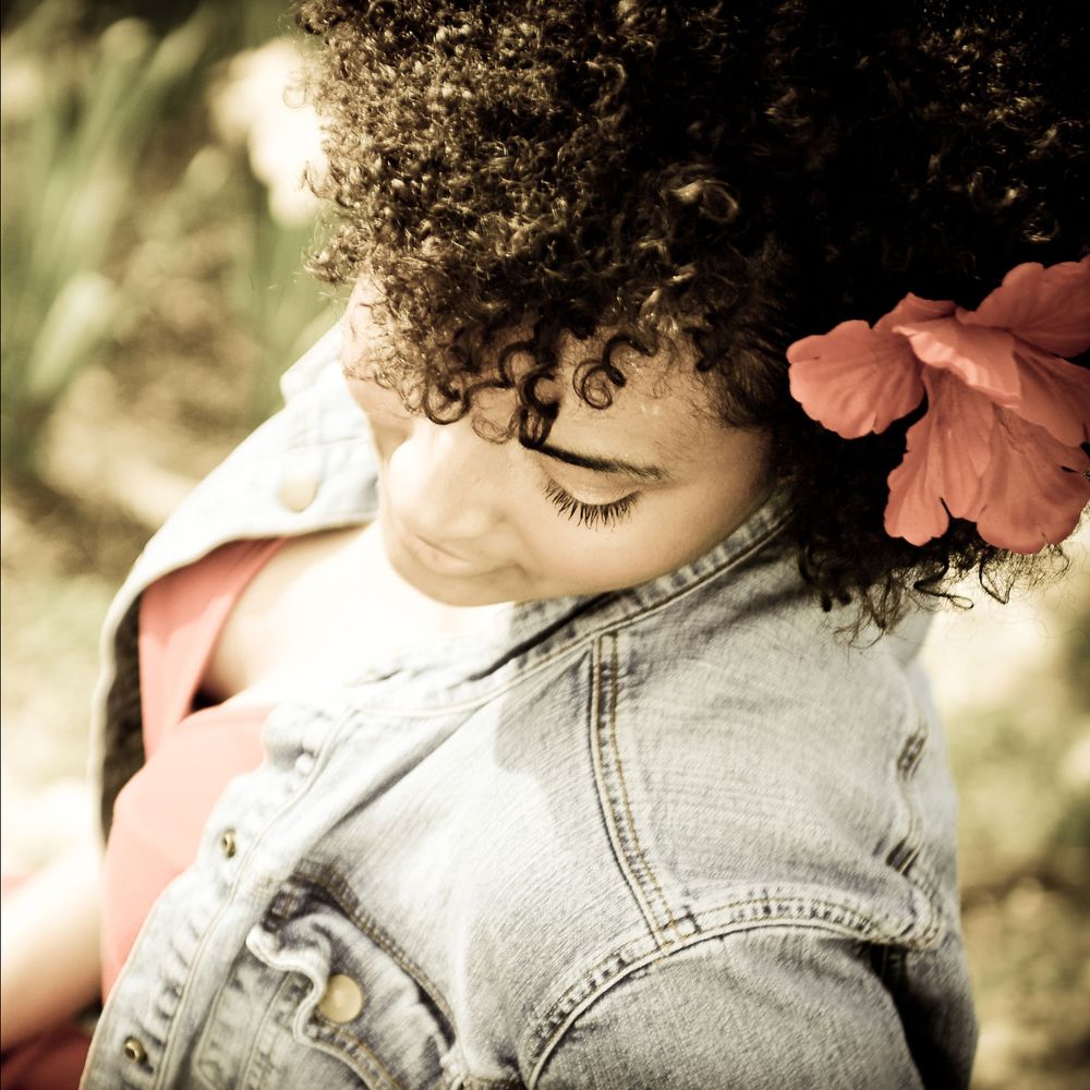 Close-up portrait of person in denim jacket with curly hair and pink flower.