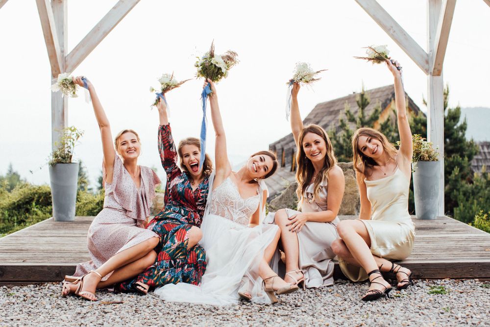 Happy bride and bridesmaids raising bouquets while sitting together at outdoor wedding venue.