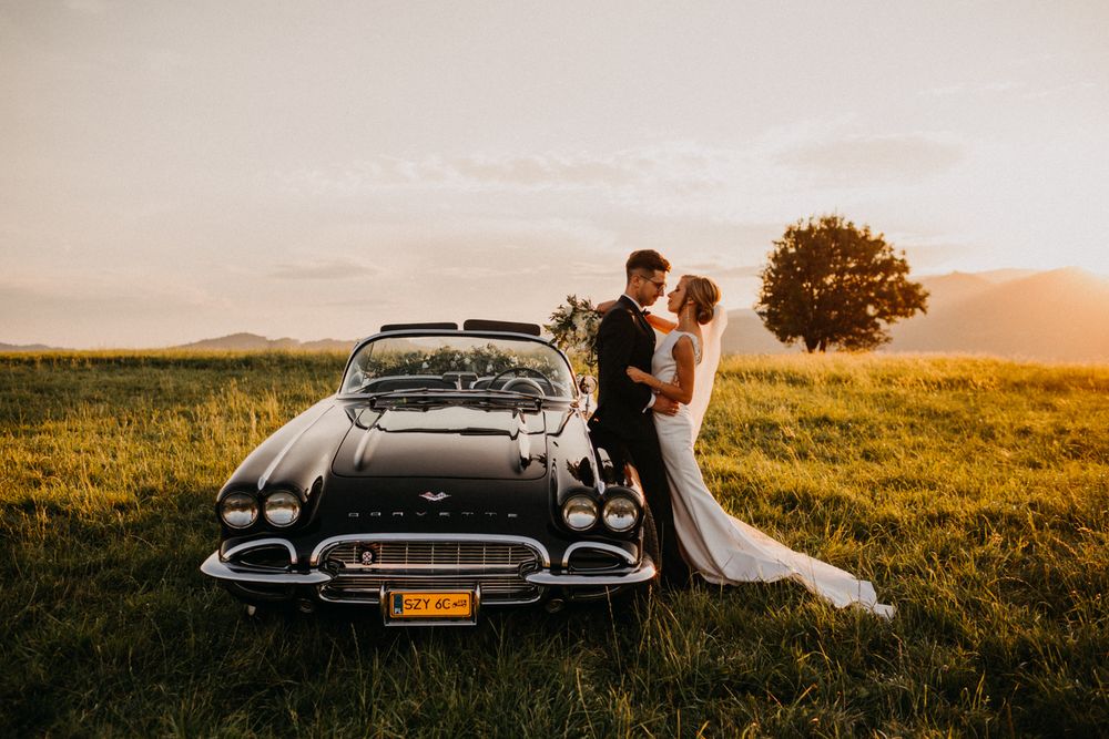 Newlyweds kissing beside vintage black Corvette in sunlit green meadow at sunset.
