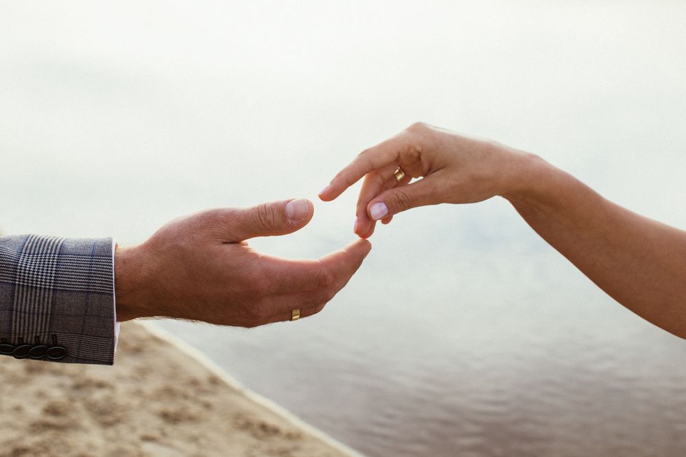 Close-up of couple exchanging wedding ring on sandy beach with water in background.