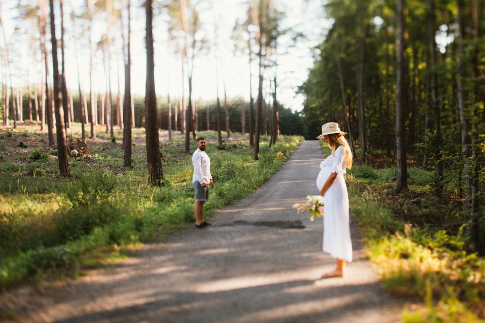 Couple standing apart on forest road, woman in white dress holding yellow flowers.