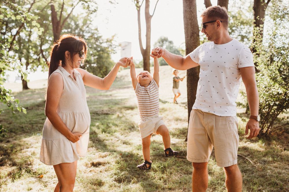 Pregnant mother and father lifting toddler by hands in sunny park setting.