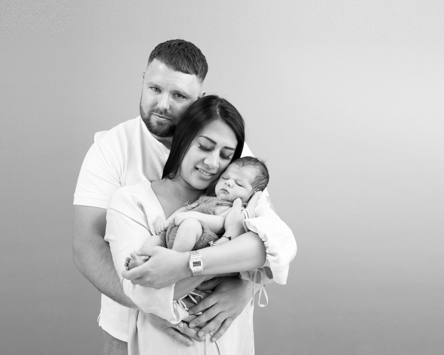 black &b white newborn family shoot, Dad has arms wrapped around mum, baby cradled in mums arms