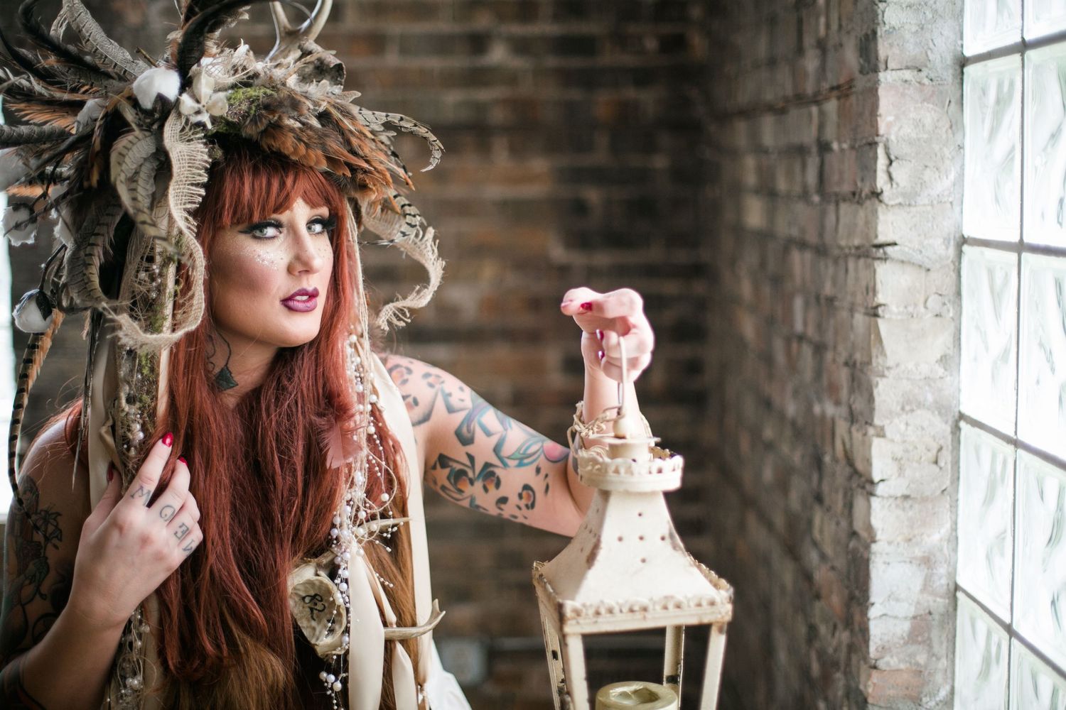 Redhead woman with tattoos and elaborate headdress holding a vintage lantern indoors.