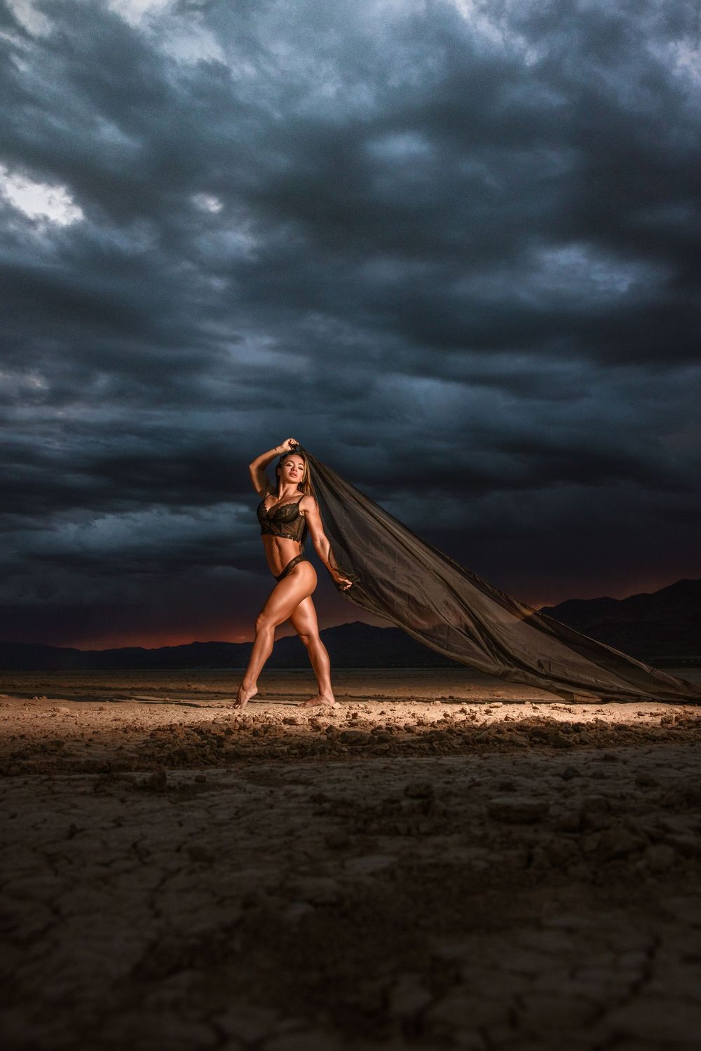Dramatic portrait of figure with flowing fabric against stormy desert sky at sunset.