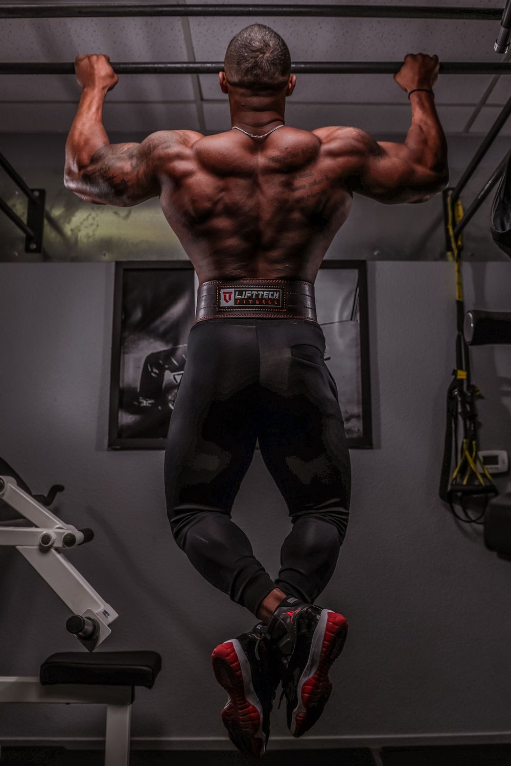 Muscular back view of athlete doing pull-ups in gym wearing black workout gear.