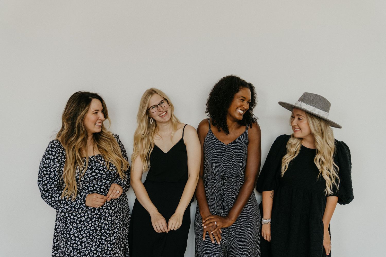Four women in black and gray outfits posing against white wall with joyful expressions.