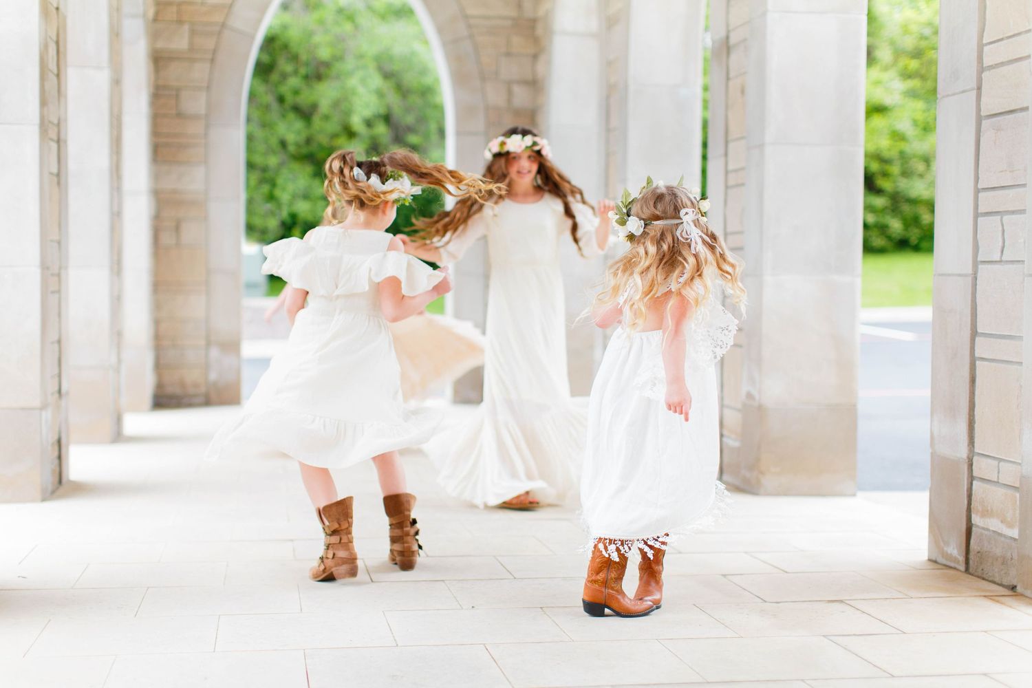 Young girls in white dresses and boots dancing in an arched stone colonnade.