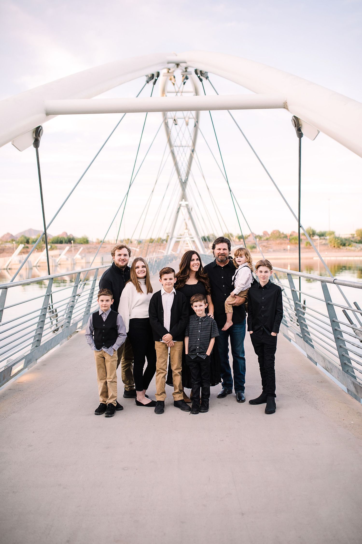 Large family of nine posing together on modern suspension pedestrian bridge at dusk.