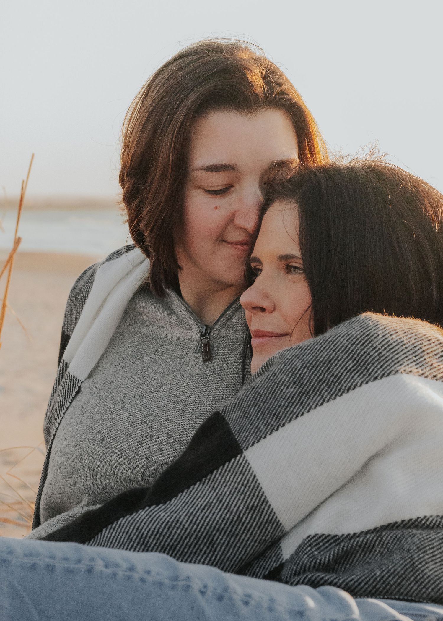 Female couple sharing intimate moments while wrapped in a cozy gray blanket on a beach at sunset.