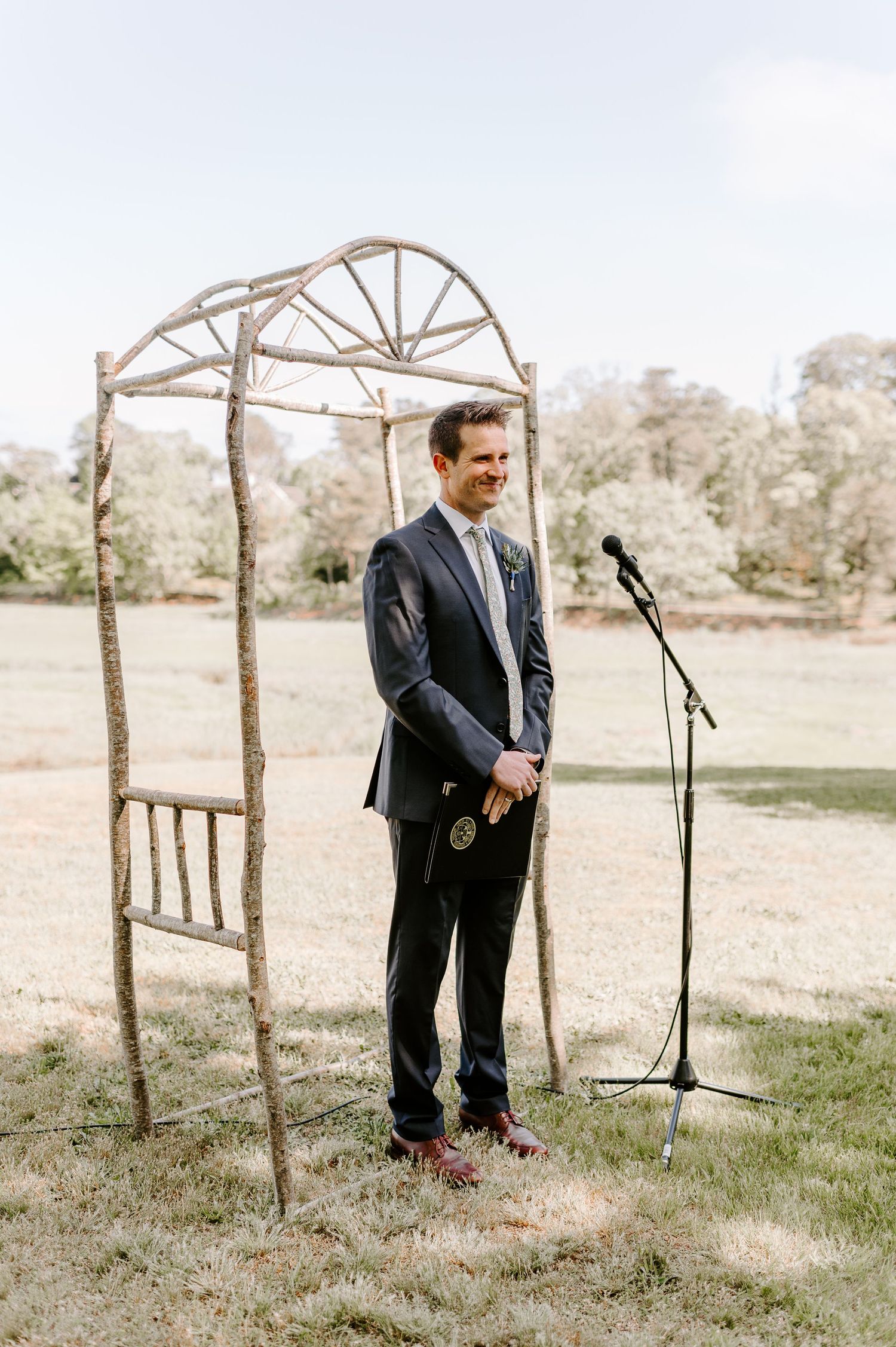 A groom in a dark suit bends down to adjust his shoe before an outdoor wedding ceremony.