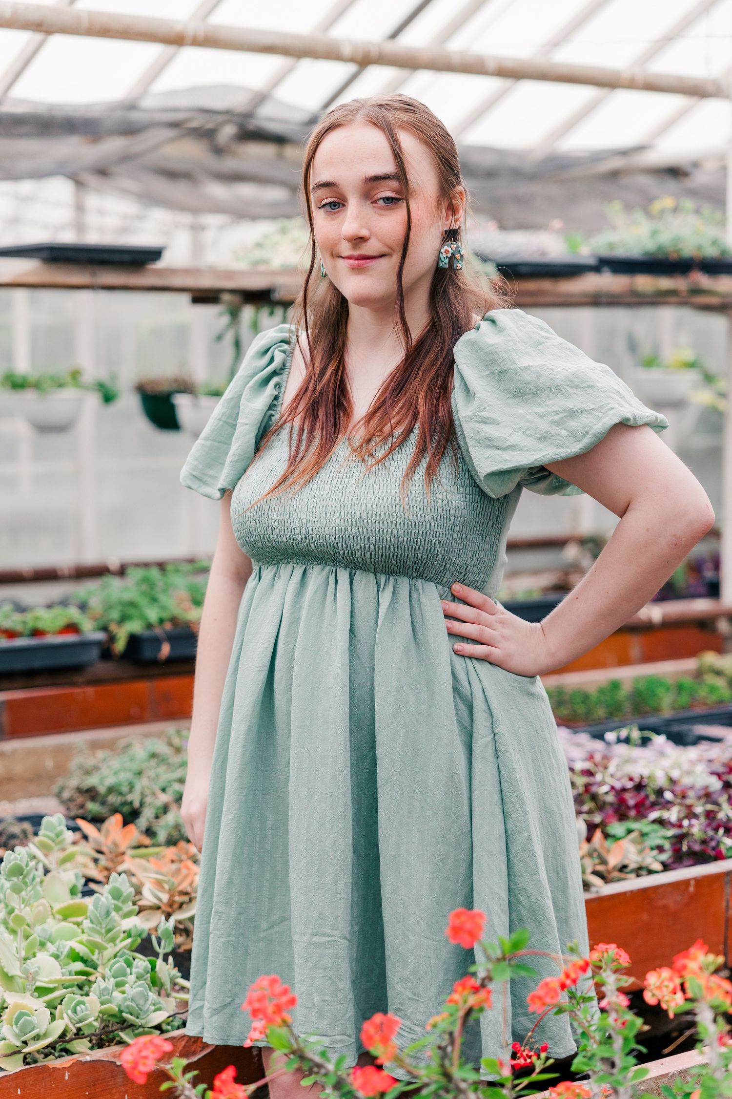 Model wearing sage green empire dress stands among potted flowers in greenhouse.