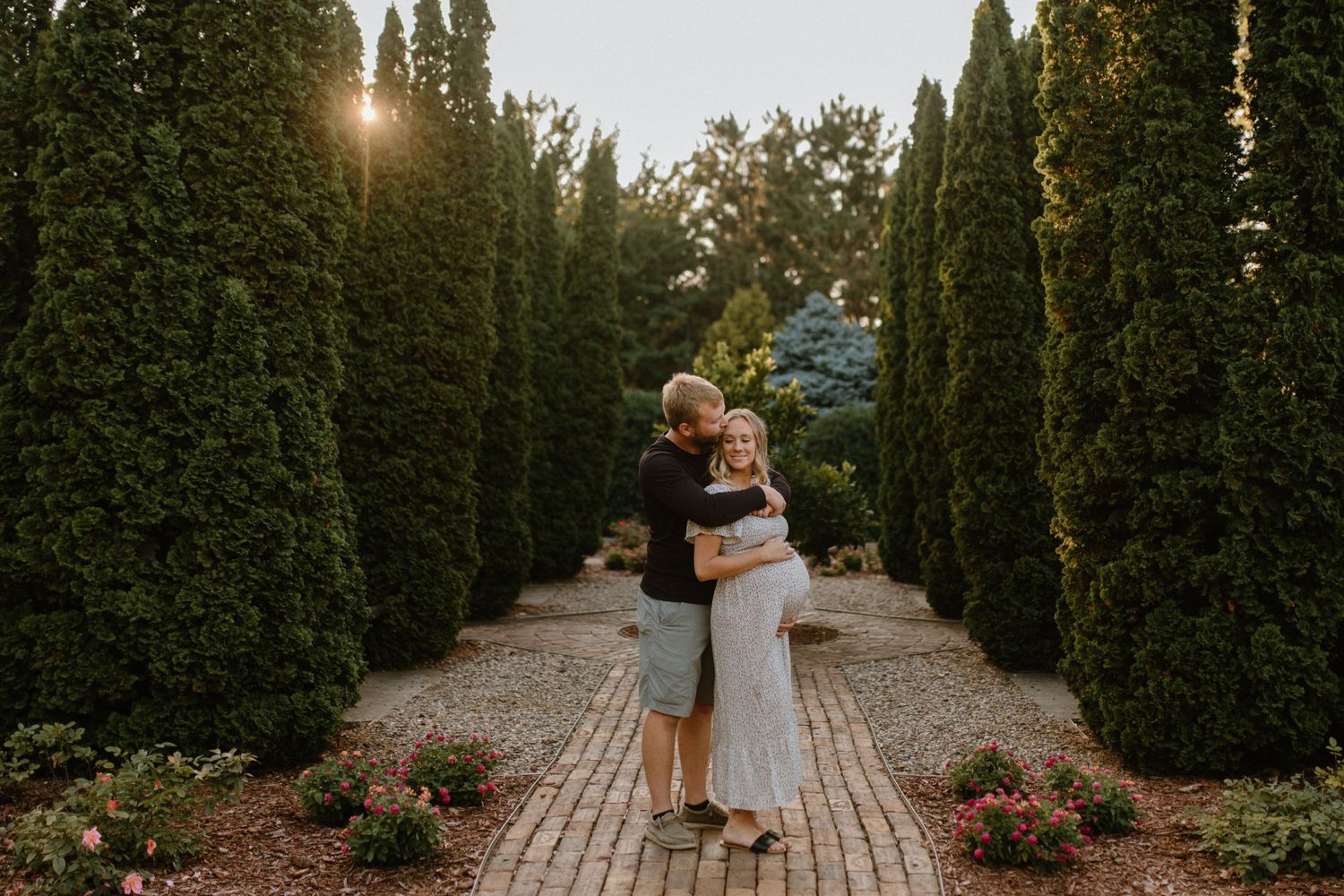 A romantic embrace between two people in a manicured garden path with cypress trees at sunset.