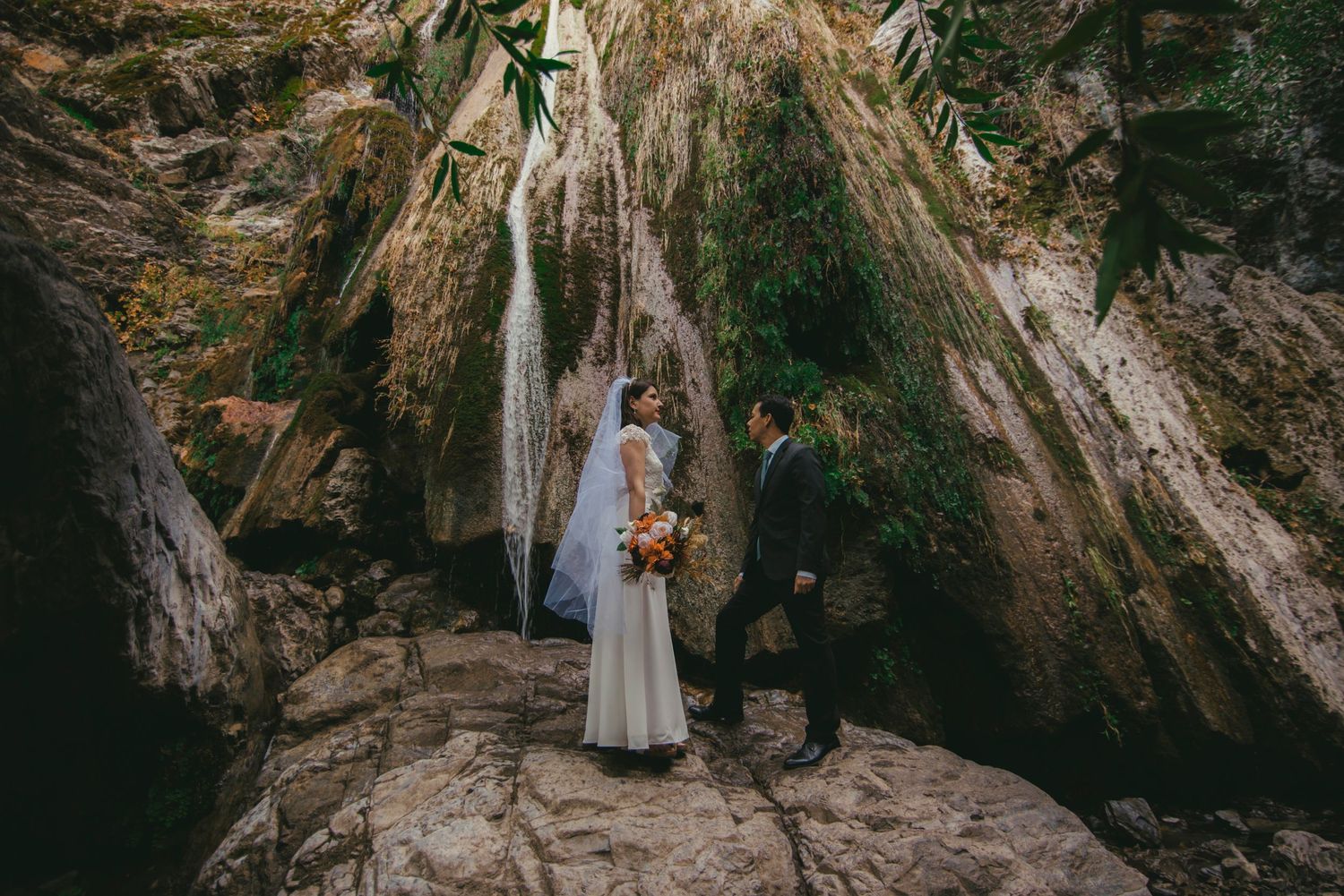A couple stands on a rocky outcrop beneath a majestic waterfall during an intimate outdoor ceremony.