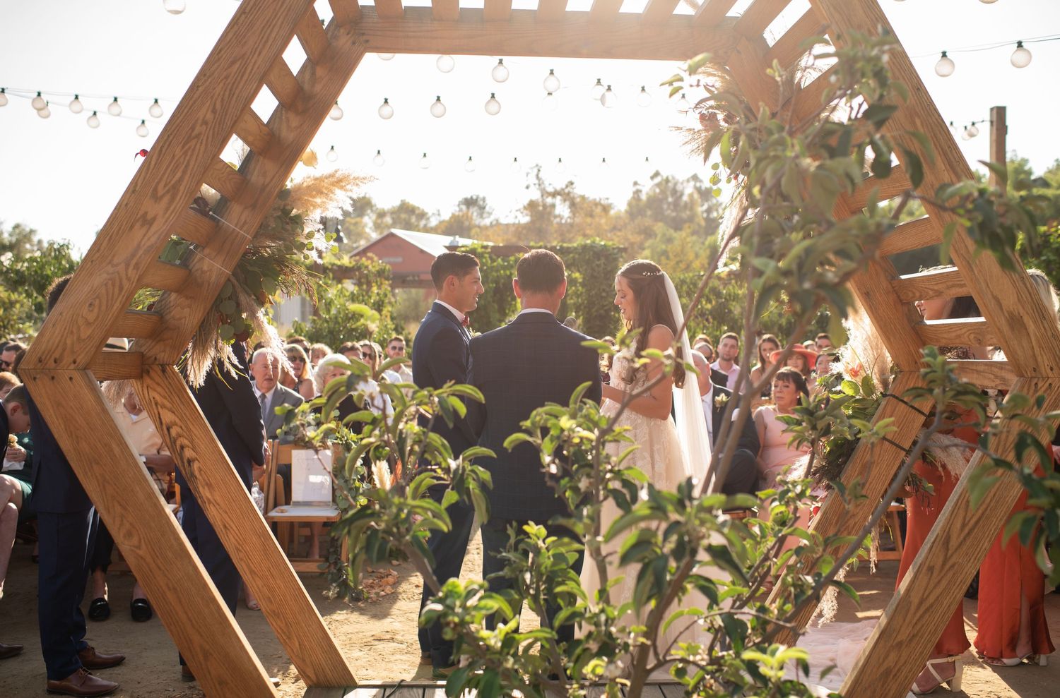 Wedding ceremony under a hexagonal wooden arch decorated with greenery and sunlit florals.