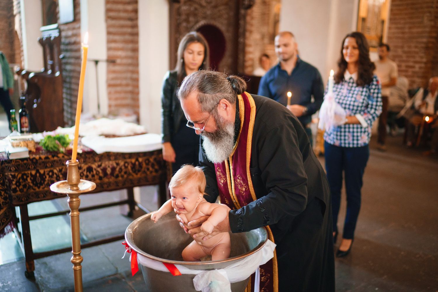 Religious baptism ceremony with priest holding baby over ornate baptismal font.