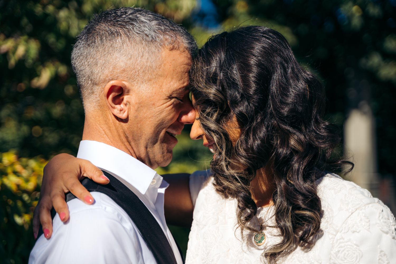 Smiling newlywed couple touching foreheads in tender romantic moment surrounded by green trees.