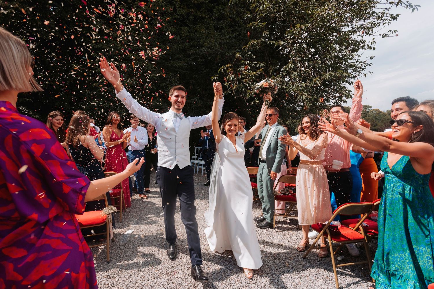 Newlyweds jump with joy while guests cheer during outdoor wedding celebration.