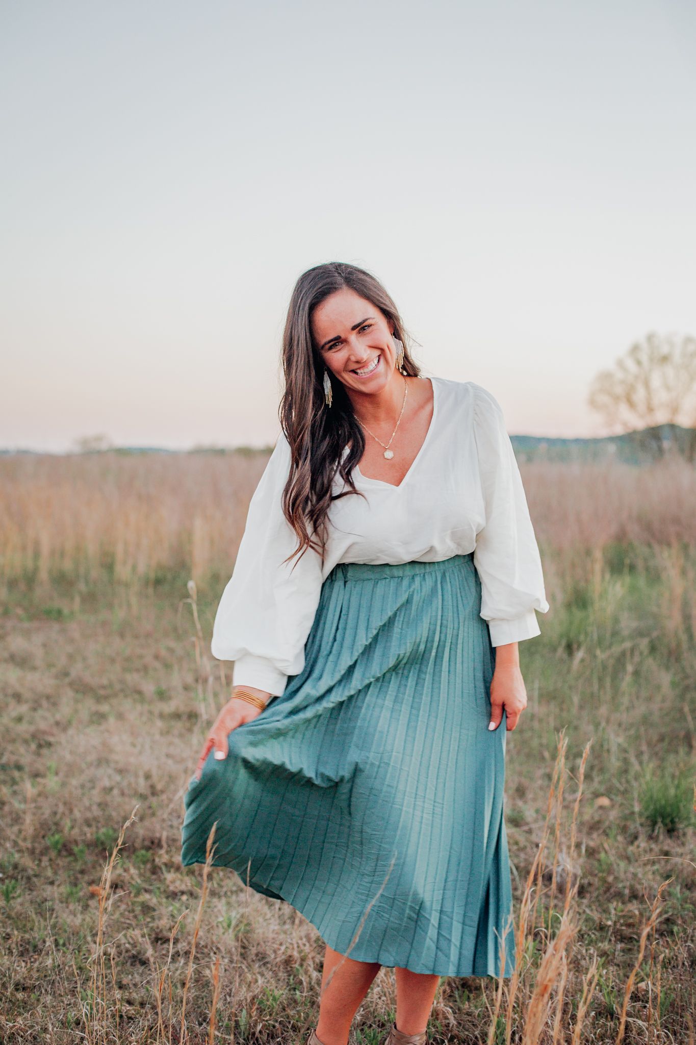 Woman in white blouse and teal pleated midi skirt poses in a golden field at sunset.