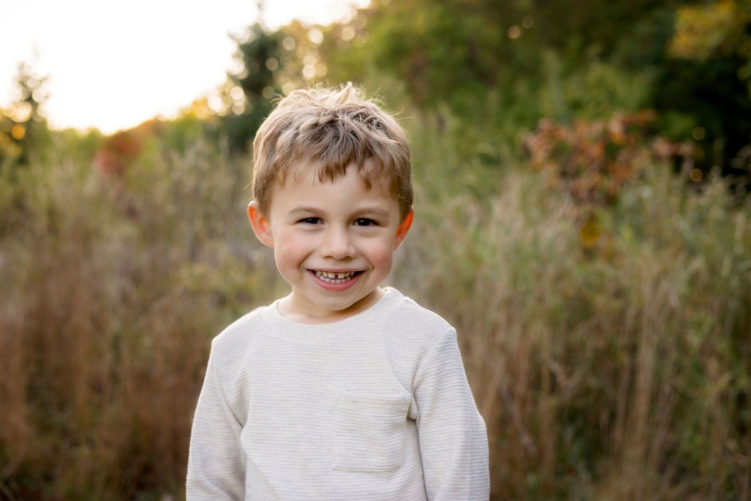 Child smiles warmly while wearing cream colored sweater outdoors.