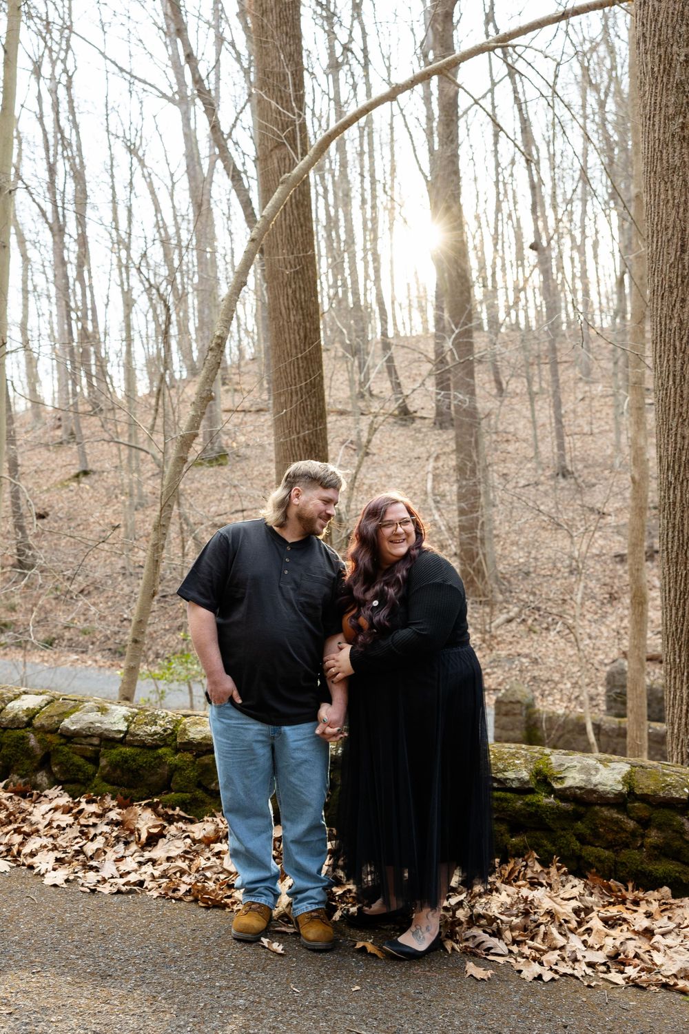 A couple poses together on a wooded path with sunlight filtering through bare winter trees.
