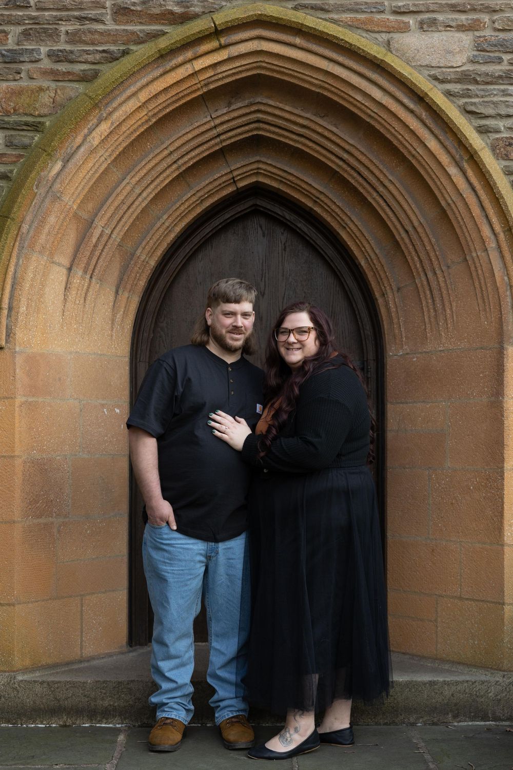 A couple poses in a gothic church doorway framed by an ornate stone arch.
