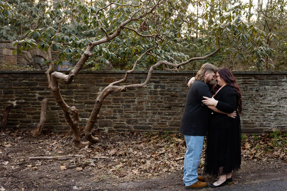 A couple embraces beside a brick wall and flowering tree in a romantic outdoor setting.