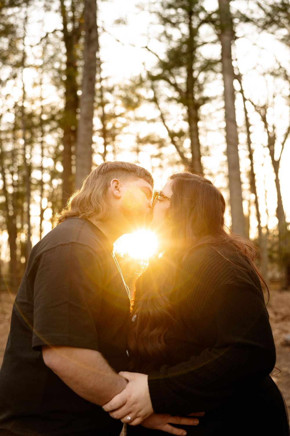 Silhouetted couple sharing a kiss with golden sunlight streaming through pine trees.