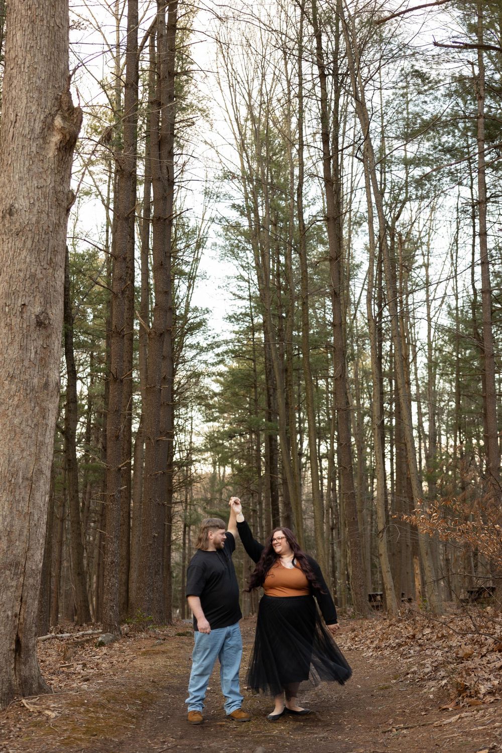 A couple walking together on a forest path surrounded by tall pine trees.