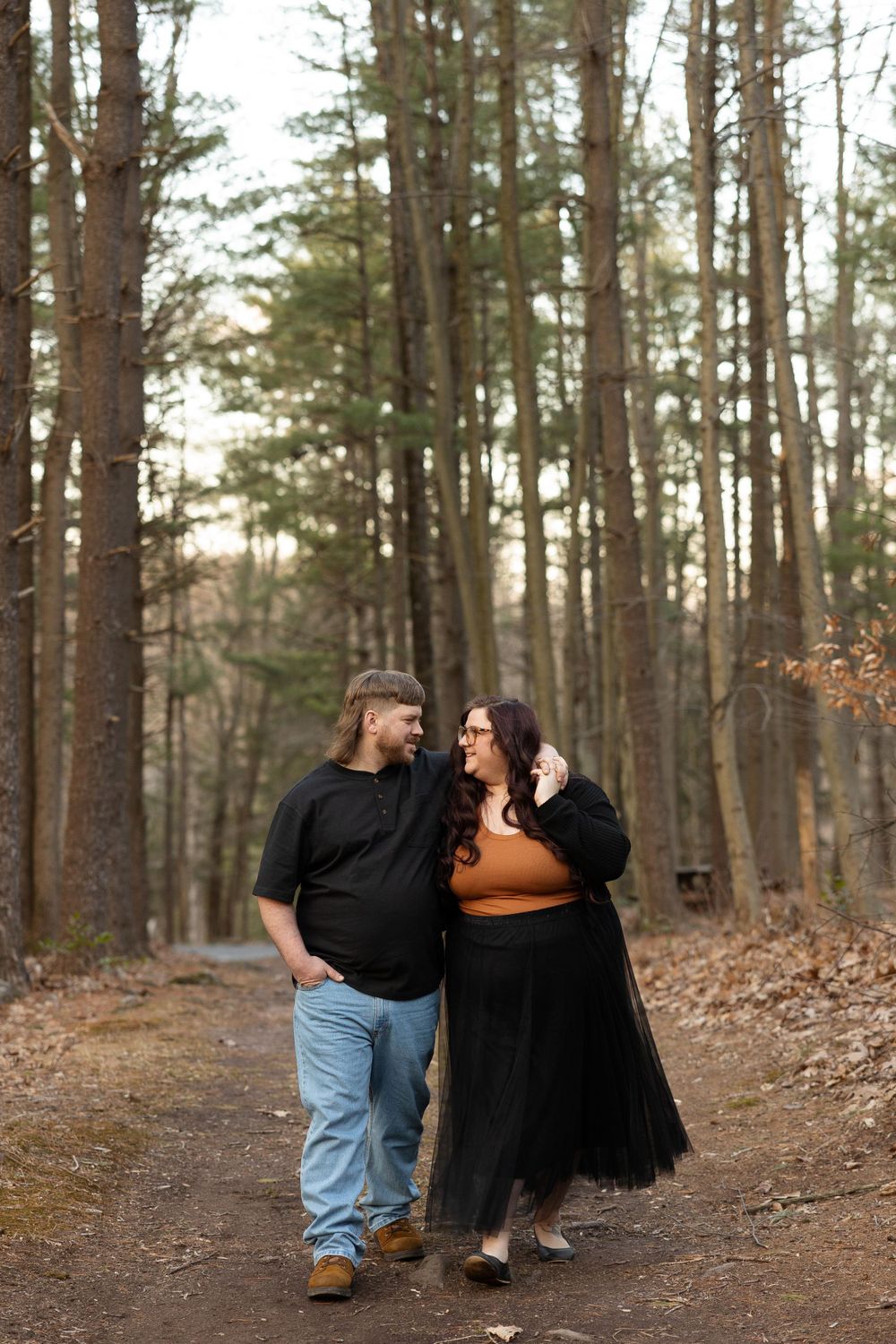 A couple takes a romantic stroll through a pine forest on a wooded trail.