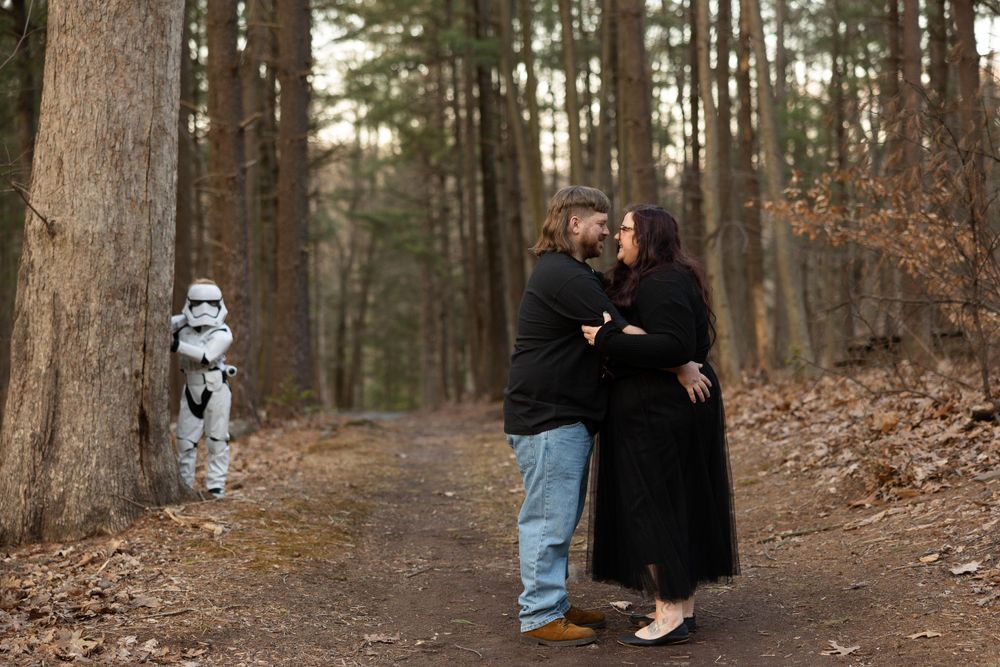 A Star Wars Stormtrooper figure follows a couple walking through a forest trail.
