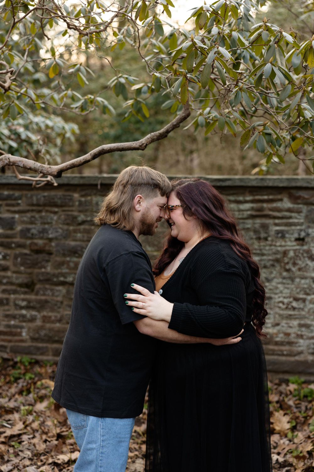A couple shares an intimate moment under green foliage against a rustic stone wall backdrop.