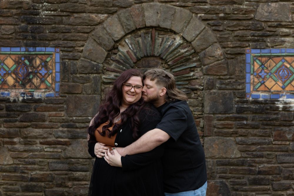 A couple embraces in front of decorative stained glass windows on a stone church wall.