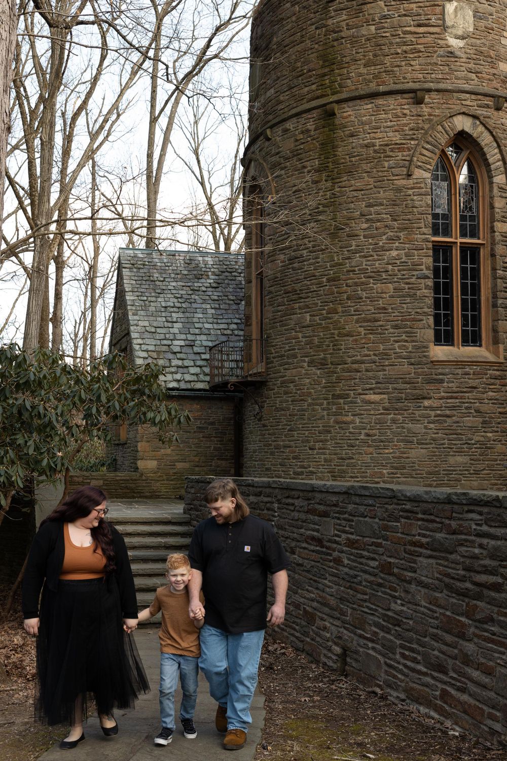 A family walks together near a stone tower with arched windows in a wooded setting.