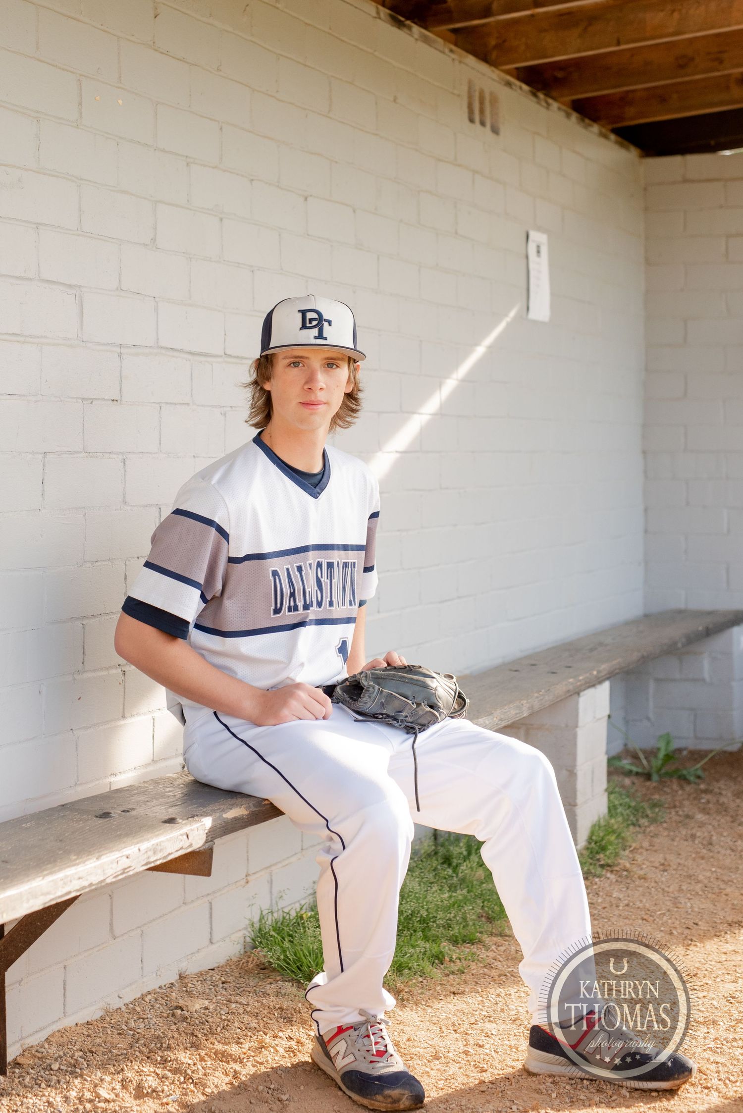 Baseball player sits on wooden bench in dugout wearing white uniform and cap.