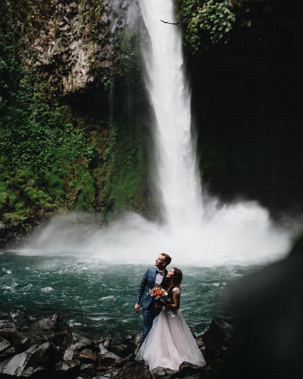 A couple stands on rocky shore beneath a powerful waterfall against mossy cliffs.
