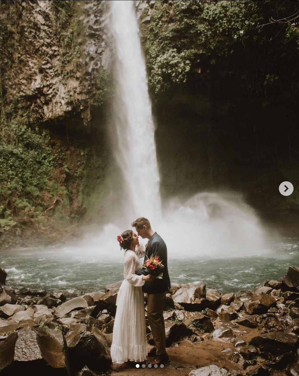 A couple shares an intimate moment before a towering waterfall with mist and rocky foreground.