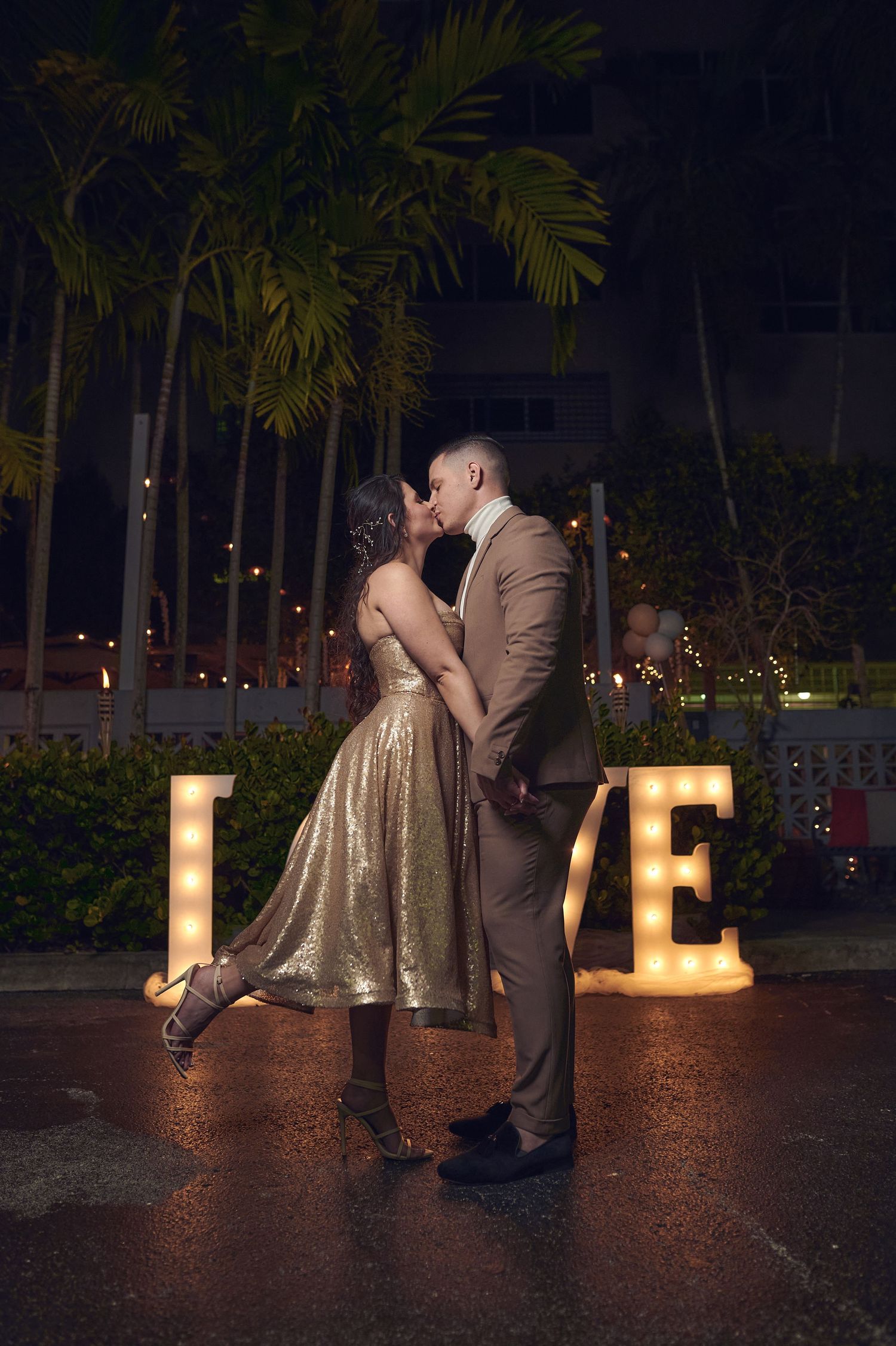Couple sharing romantic kiss near illuminated LOVE sign with palm trees at night.
