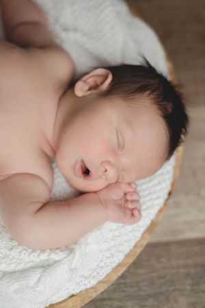Peaceful newborn baby sleeping with mouth open on soft white blanket in wooden bowl.