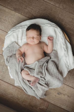 Newborn baby sleeping in round wooden bowl with white and gray knit blankets.