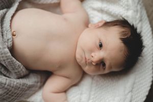 Alert dark-haired newborn baby lying on white blanket wrapped in gray muslin cloth.