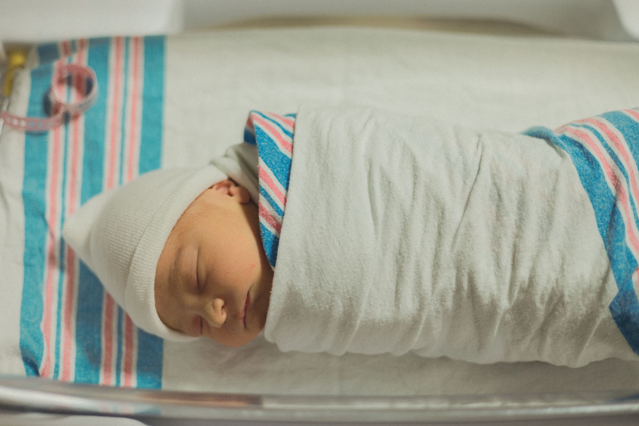 Newborn baby swaddled in white blanket with striped hospital hat sleeping in bassinet.