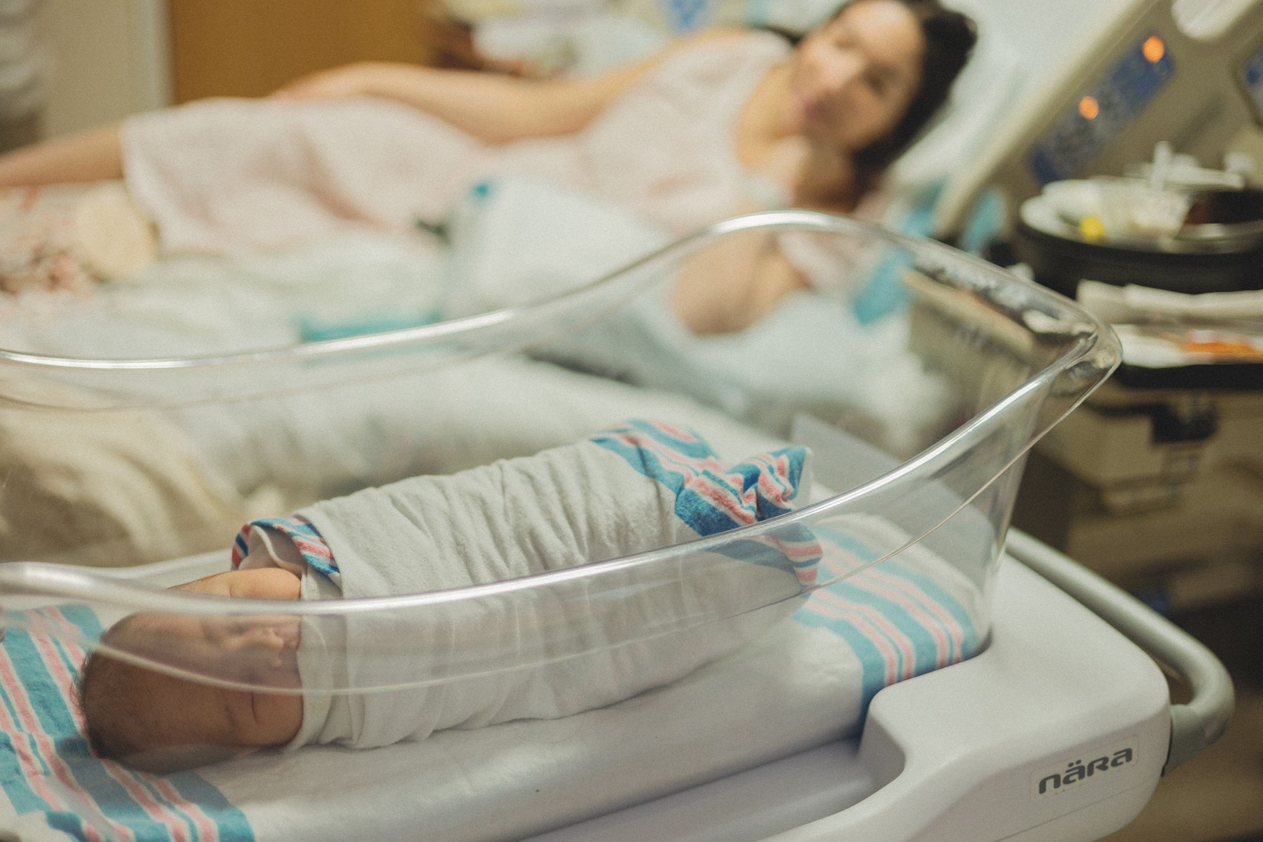 Swaddled newborn in hospital bassinet with mother resting in delivery room bed behind.
