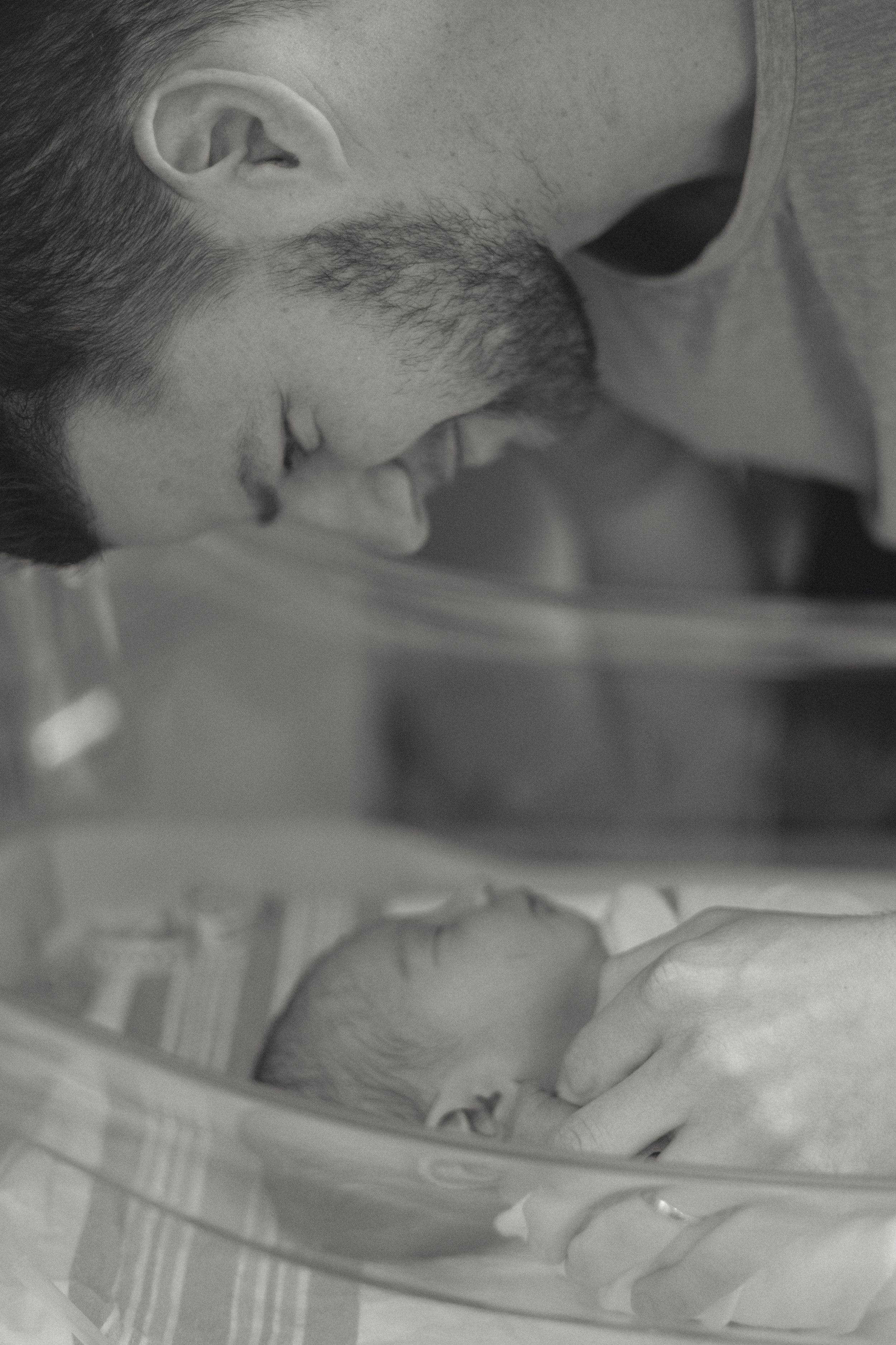 Black and white photo of a father leaning over to kiss his newborn baby in a hospital bassinet.
