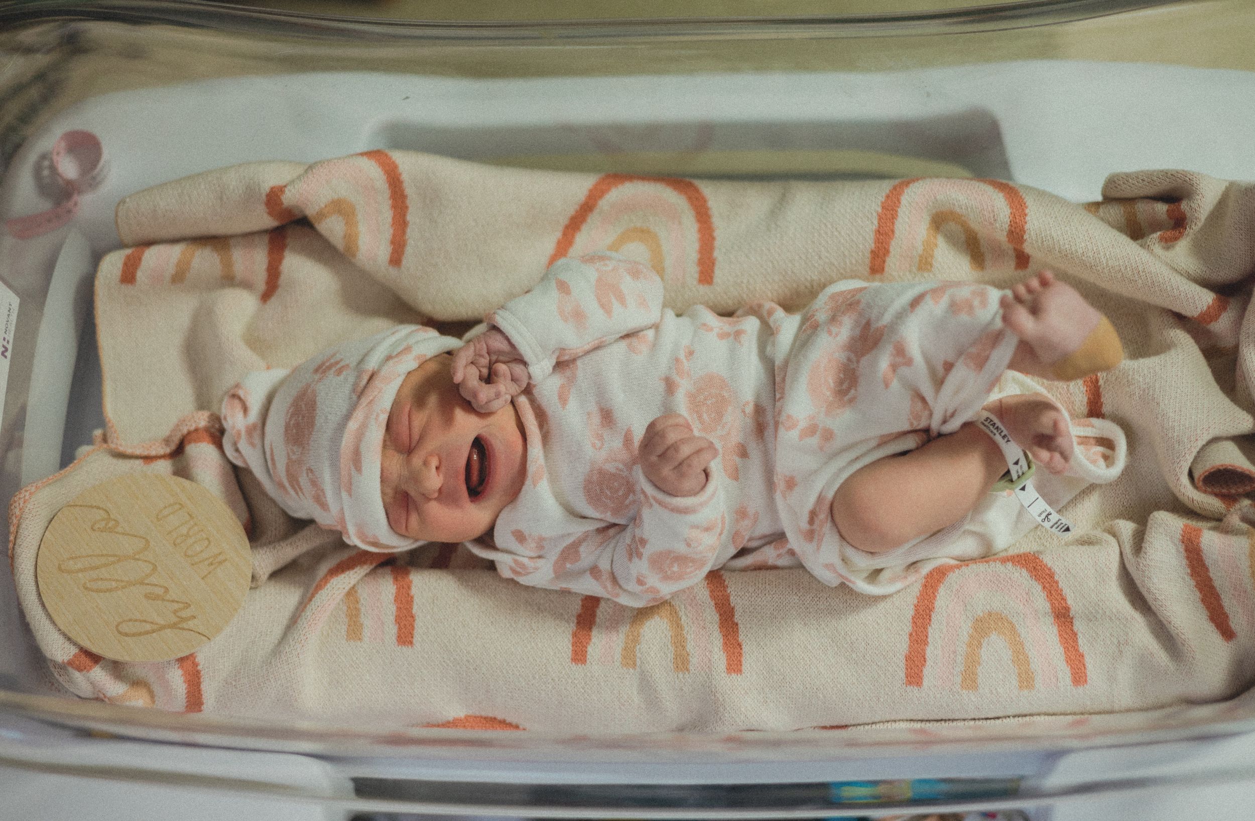 Crying newborn baby in rainbow print outfit lying in hospital bassinet with wooden name tag.