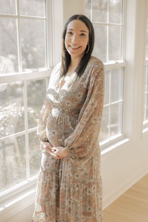 Smiling pregnant woman in floral maternity dress cradling baby bump by bright windows.
