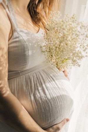 Pregnant woman in grey dress holding baby's breath flowers beside sunlit window.