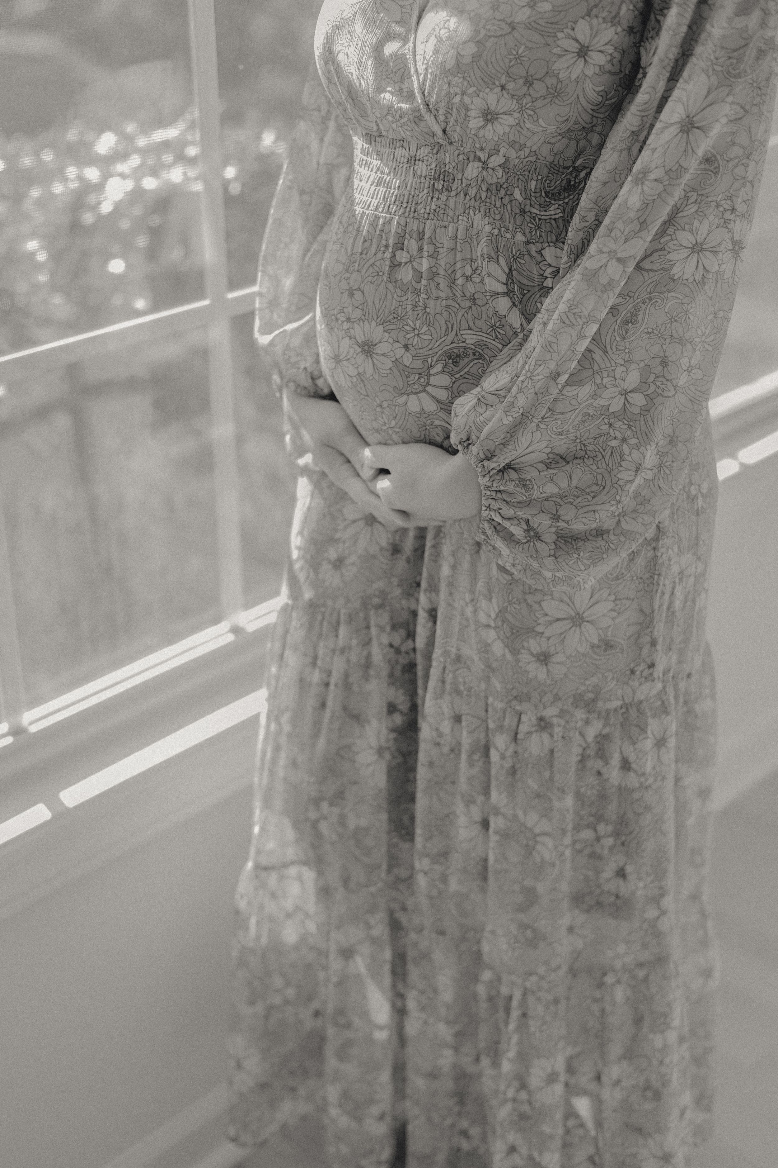 Black and white photo of pregnant woman in floral dress cradling baby bump by window.